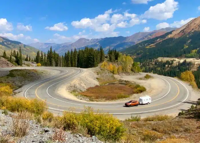 orange jeep tows white camper around curvy colorado mountain road with trees and mountains