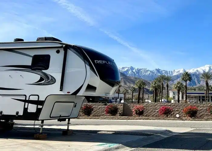 fifth wheel trailer parked in campsite red bushes palm trees snow capped mountains behind