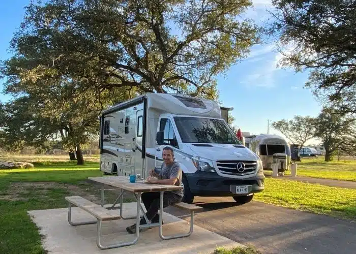 man sits at picnic table by rv in campsite trees and grass